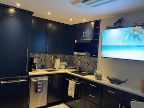 A modern kitchen with dark cabinetry, a built-in oven, a microwave, and a large flat-screen TV mounted on the wall. - Waterfront Condo-fully renovated, in the desirable West End, steps to the beach. (Provincetown)