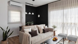 A modern living room with a beige sofa, a coffee table with fruit, a potted plant, and a black accent wall. - AirTLV - Arlozorov Apartment's (Ramat Gan)