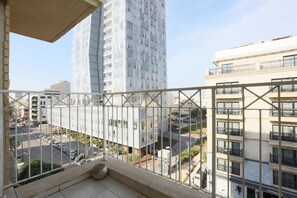 Un balcon offrant une vue sur le paysage urbain, avec notamment un immeuble de grande hauteur et une rue bordée de voitures garées.