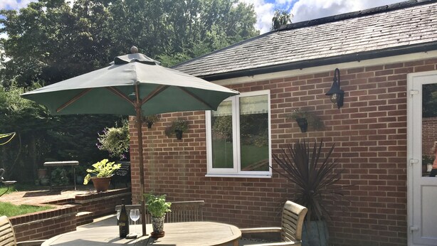 An outdoor patio with a round table and wooden chairs, a green umbrella, and potted plants. - Charming 1-bedroom Apartment Near Hailsham (Hailsham)