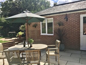 An outdoor patio with a round table and wooden chairs, a green umbrella, and potted plants. - Charming 1-bedroom Apartment Near Hailsham (Hailsham)