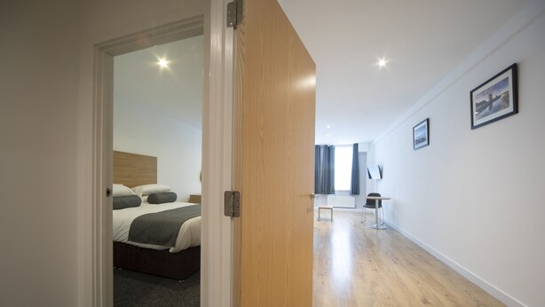A modern hotel room with a wooden door leading to a bedroom with a bed and a desk. - Wellington Apartments (Norwich)