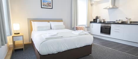 A modern hotel room with a neatly made bed, a small bedside table with a lamp, a kitchenette with a stove and toaster, and a large window with curtains.