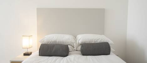 A neatly made bed with white linens and two gray pillows, a bedside table with a lamp, and a plain white wall.