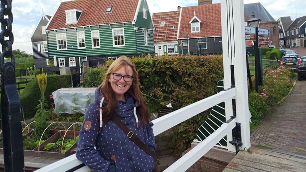 Een persoon zit op een witte metalen reling met uitzicht op een straat met traditionele huizen en een kerktoren.
