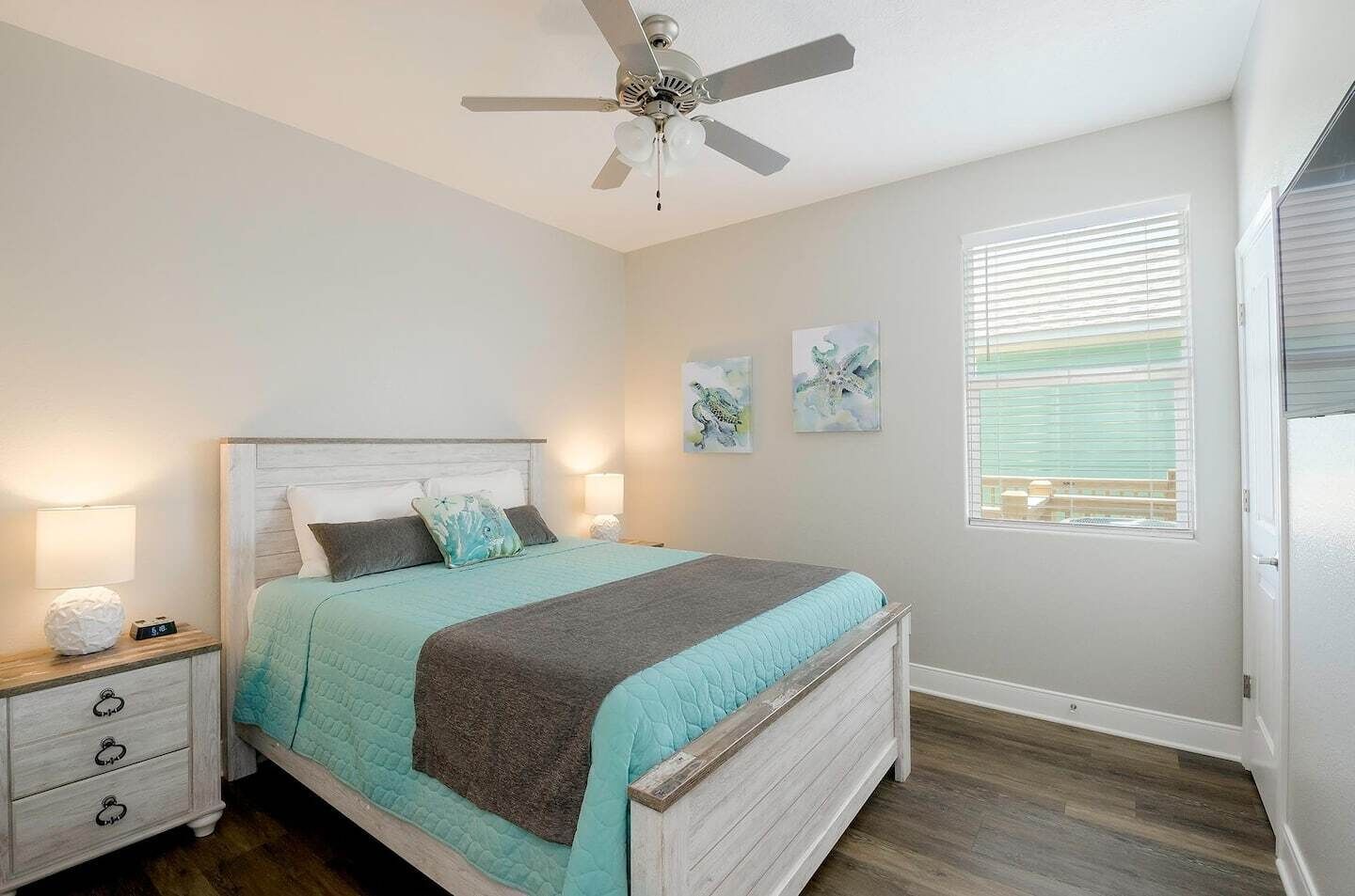 Une chambre à coucher comprenant un lit, des tables de chevet, un ventilateur de plafond, des œuvres d’art accrochées au mur et une fenêtre avec des stores.