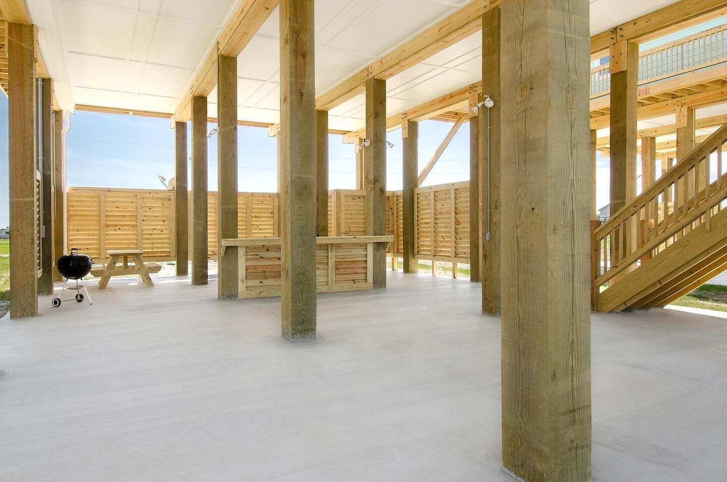 A spacious covered area with wooden beams and a concrete floor, featuring a barbecue grill and picnic table.