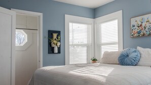 A bedroom with a large bed, white walls, and a window with blinds. - Charming cozy cottage on historic numbered streets (Idaho Falls)