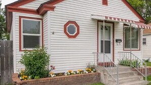 A single-story house with a red roof, white siding, and a brick flower bed in front. - Charming cozy cottage on historic numbered streets (Idaho Falls)