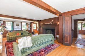 A spacious living room with a green sofa, a fireplace, wooden beams, and large windows.