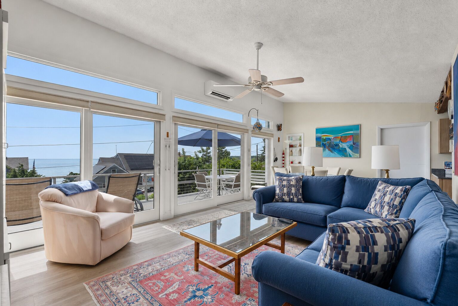 A spacious living room with a blue sofa, a beige armchair, a glass coffee table, and a ceiling fan.