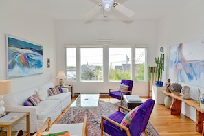 A spacious living room with a white sofa, a glass coffee table, and a purple armchair. There are large windows providing a view of the outside.