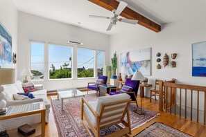 A spacious living room with a large window, a ceiling fan, and a view of the sea.
