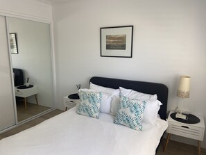 A neatly made bed with white bedding and two decorative pillows, a bedside table with a lamp, and a framed picture on the wall.