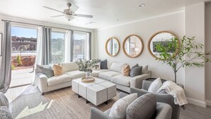 A modern living room with a sectional sofa, a coffee table, and a large window with curtains. There are two round mirrors on the wall and a ceiling fan.