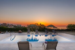 A swimming pool with lounge chairs and umbrellas, surrounded by a landscaped area with a sunset view. - Villa Chrysalida, peaceful home with scenic views! (Skaleta)