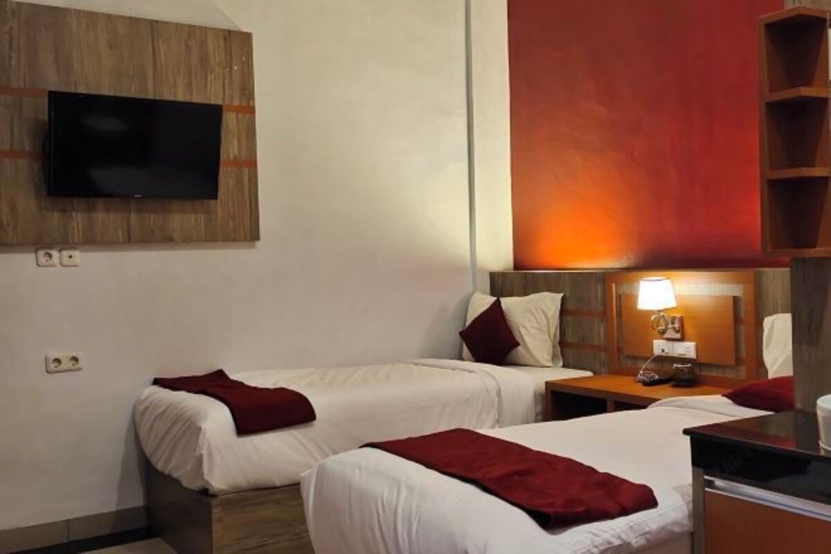 A hotel room with two beds, a TV mounted on a wooden panel, a nightstand with a lamp, and a wooden shelf unit.