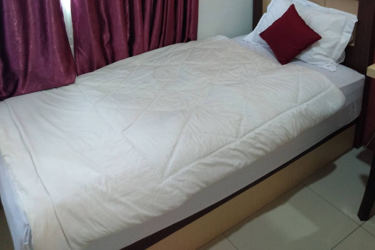 Un lit simple avec un matelas blanc et un oreiller rouge, une tête de lit en bois et un rideau rouge tiré partiellement sur le côté.