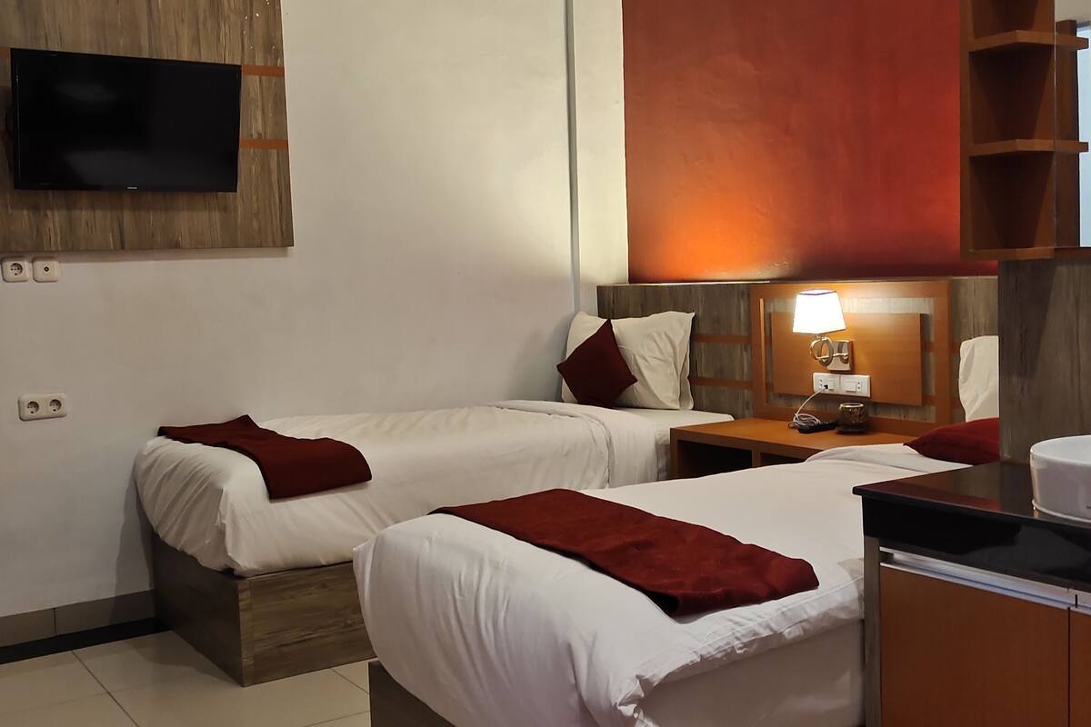 Une chambre d’hôtel avec deux lits, un téléviseur fixé au mur, une table de chevet avec une lampe, un lavabo et un miroir.