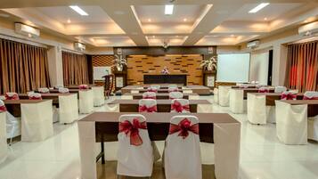Un salón de conferencias con filas de mesas y sillas, un escenario con fondo de madera y una pantalla de proyector.