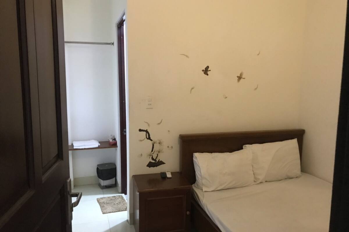 Ein Schlafzimmer mit einem Bett, Nachttisch und einem kleinen Beistelltisch. Es gibt ein Regal mit einem Handtuch und einem Mülleimer.