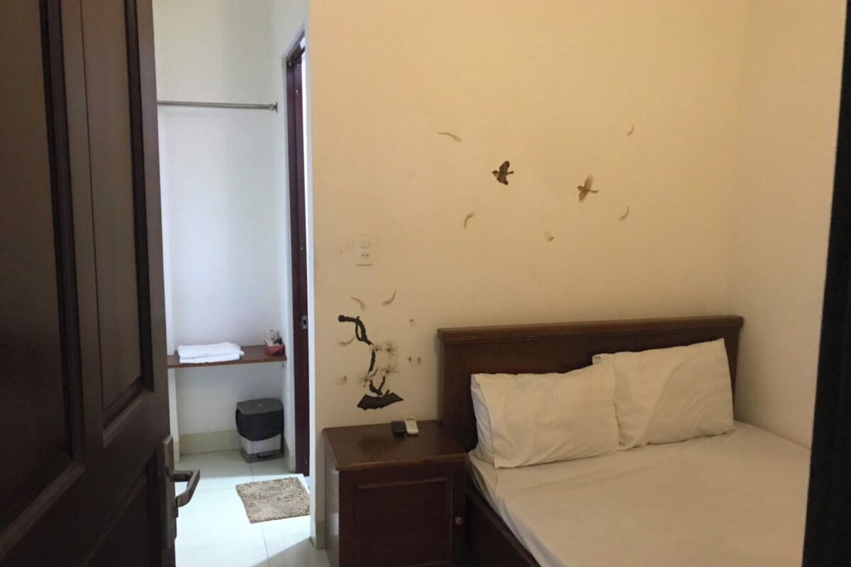 A bedroom with a wooden bed, a bedside table, a nightstand, a small shelf, a trash bin, and a door.