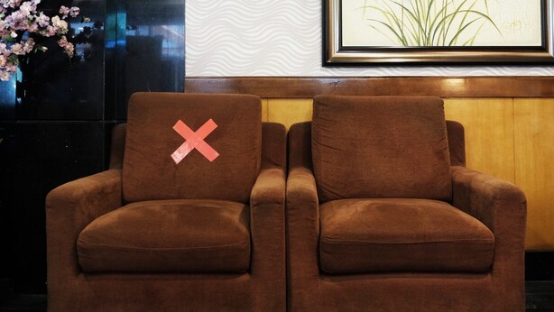 A brown sofa with a red 'X' on one cushion, positioned against a wall with a framed picture and a vase of flowers.
