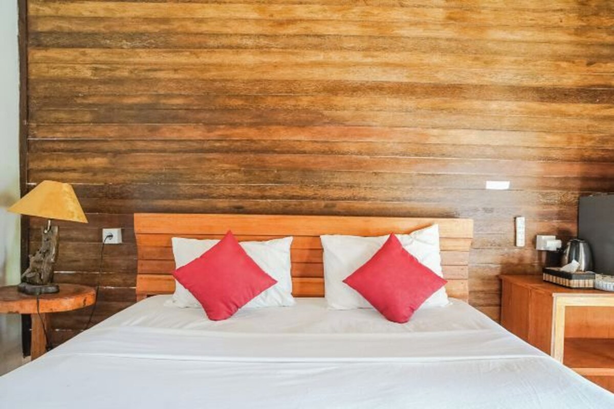 A bed with white bedding and red pillows, a wooden headboard, a nightstand with a lamp, and a wooden wall.