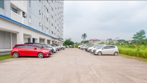 A parking lot with cars parked in rows, a building with multiple floors and balconies, and greenery in the background. - Room Rendi @ Apt Dramaga Tower (Dramaga)