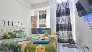 A bedroom with a bed, a desk, a chair, a TV, and a window with curtains. - Room Rendi @ Apt Dramaga Tower (Dramaga)