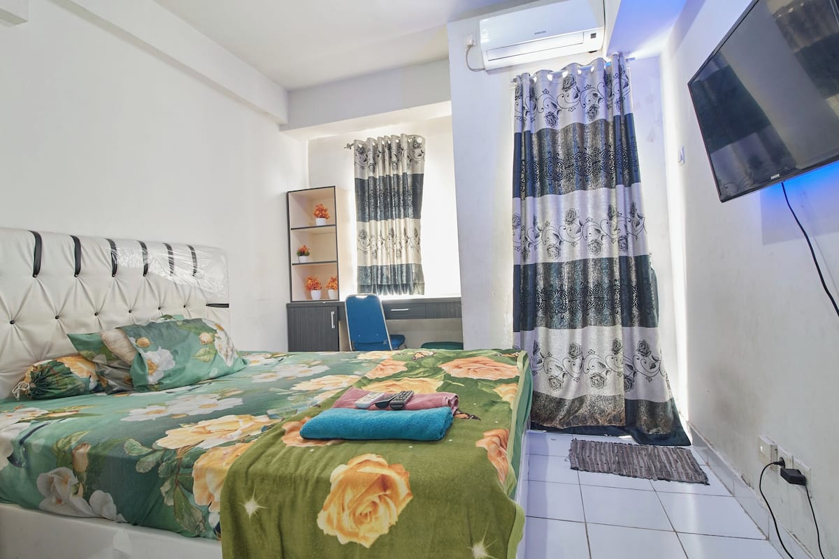 A bedroom with a bed, a desk, a chair, a TV, and a window with curtains.