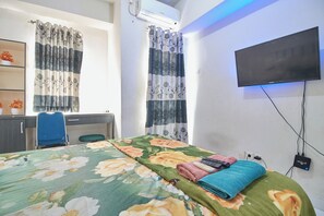 A bedroom with a bed, a desk, a chair, a TV, and a shelf with flowers. - Room Rendi @ Apt Dramaga Tower (Dramaga)