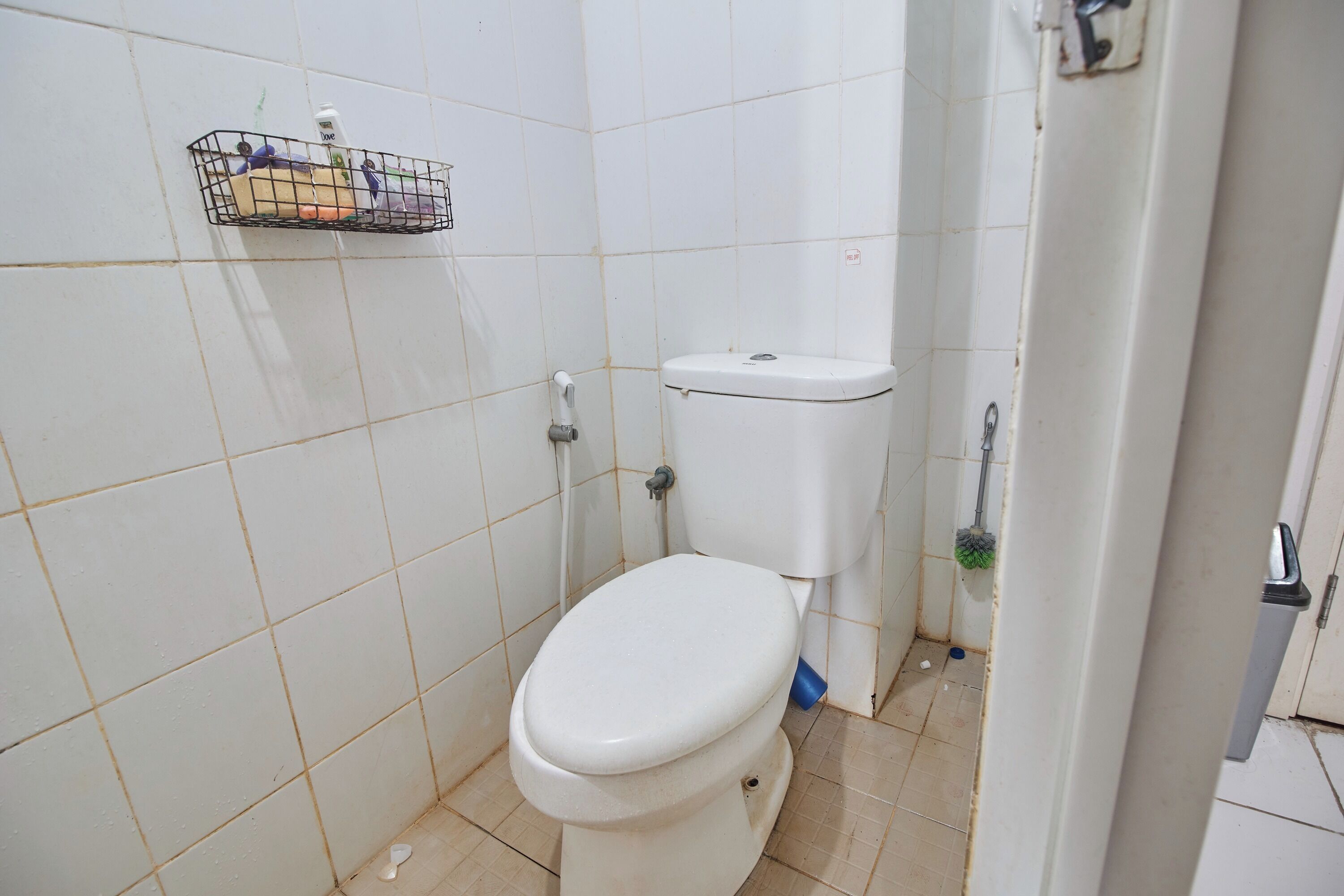 Pieni, siisti kylpyhuone, jossa on wc, seinälle kiinnitetty hylly, jossa on hygieniatarvikkeita, ja lattialla seinää vasten nojaava moppi.