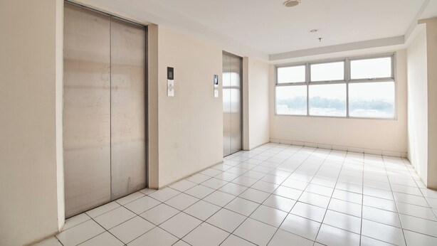 A spacious, empty room with white tiled floors and walls, two elevators, and large windows with a view of the sky. - Room Rendi @ Apt Dramaga Tower (Dramaga)