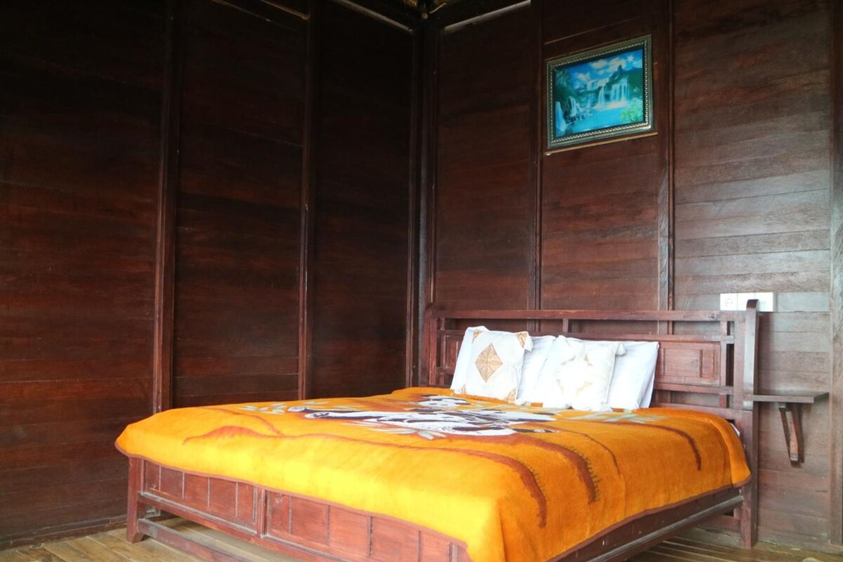 A wooden bed with a headboard, a framed picture on the wall, and a woven mat on the floor.