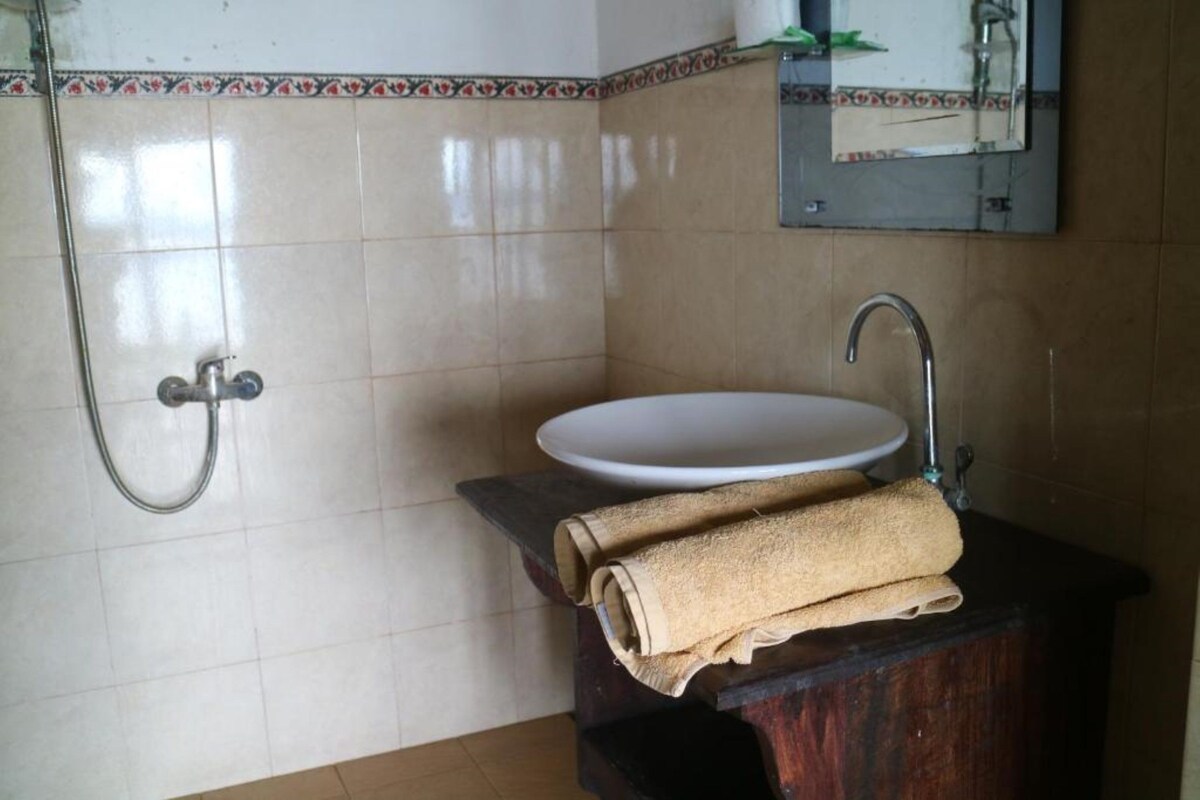 Un baño con lavamanos blanco, tocador de madera, una toalla y ducha con manguera metálica.