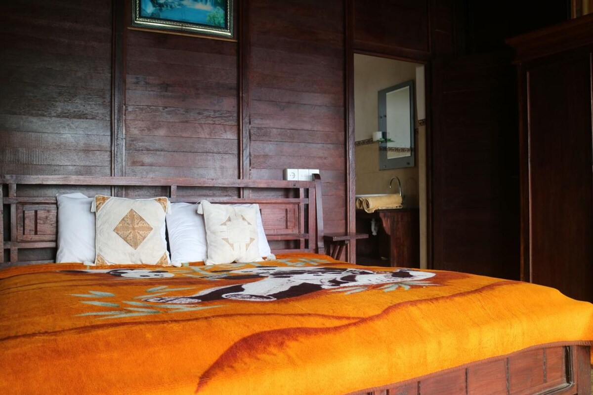 A wooden bed with a patterned blanket and pillows, a wooden headboard, and a view into a bathroom.
