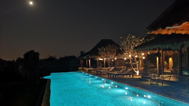 Un área de piscina nocturna con sillones de descanso y sombrillas, rodeada de árboles y una montaña al fondo.