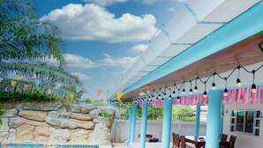 Un espacio al aire libre techado con piscina, pared de piedra y asientos bajo un techo azul con luces colgantes.