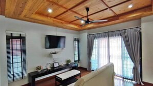 A living room with a wooden ceiling, a flat-screen TV, a sofa, a coffee table, and a window with curtains. - Lan House Pool Villa (Chiang Mai)