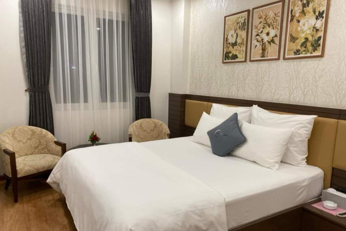 Ein Hotelzimmer mit einem Bett, zwei Sesseln, einem Nachttisch mit Telefon und einem Fenster mit durchsichtigen Vorhängen.