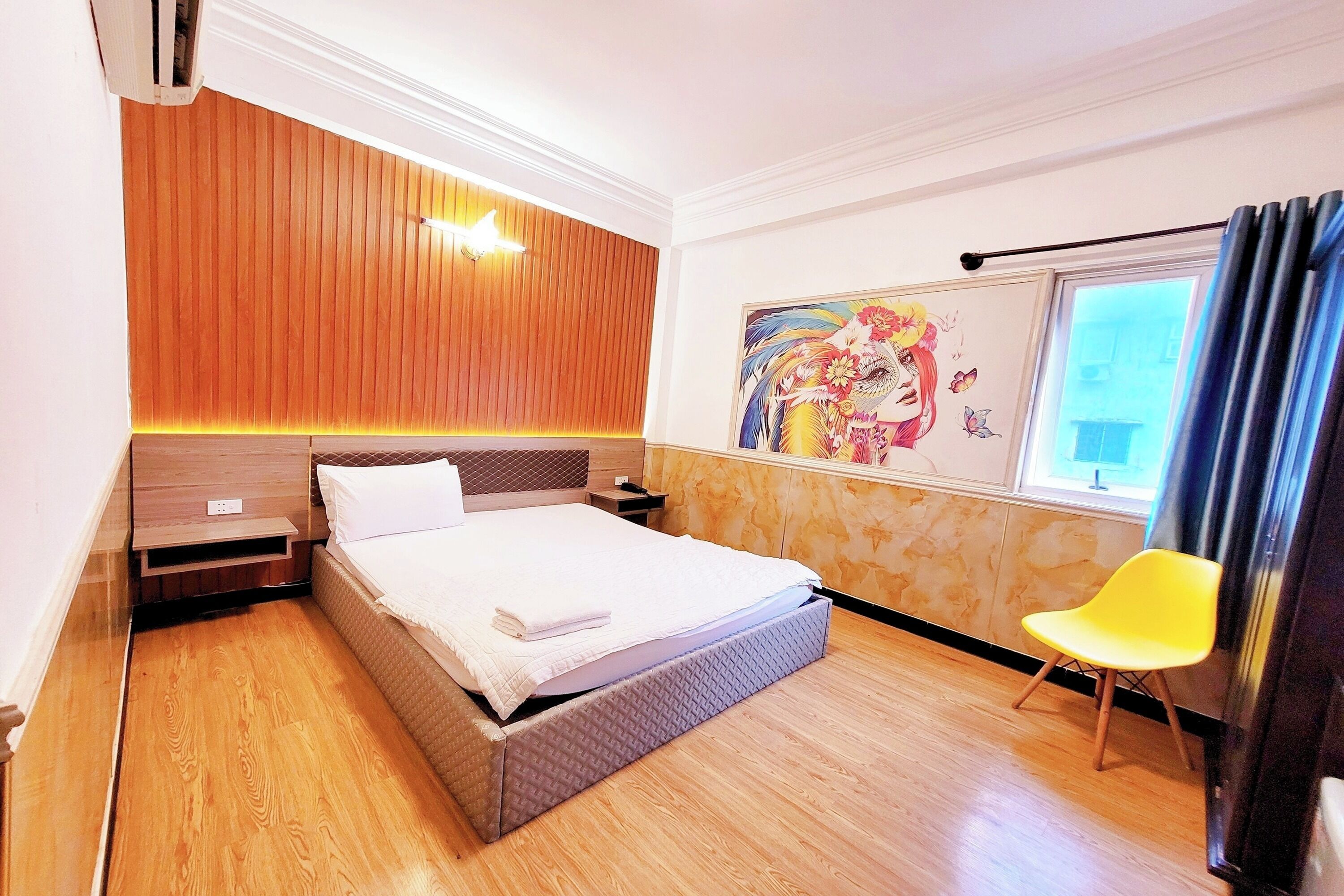 A hotel room with a wooden panel wall, a bed with a white comforter, a yellow chair, and a large window with curtains.