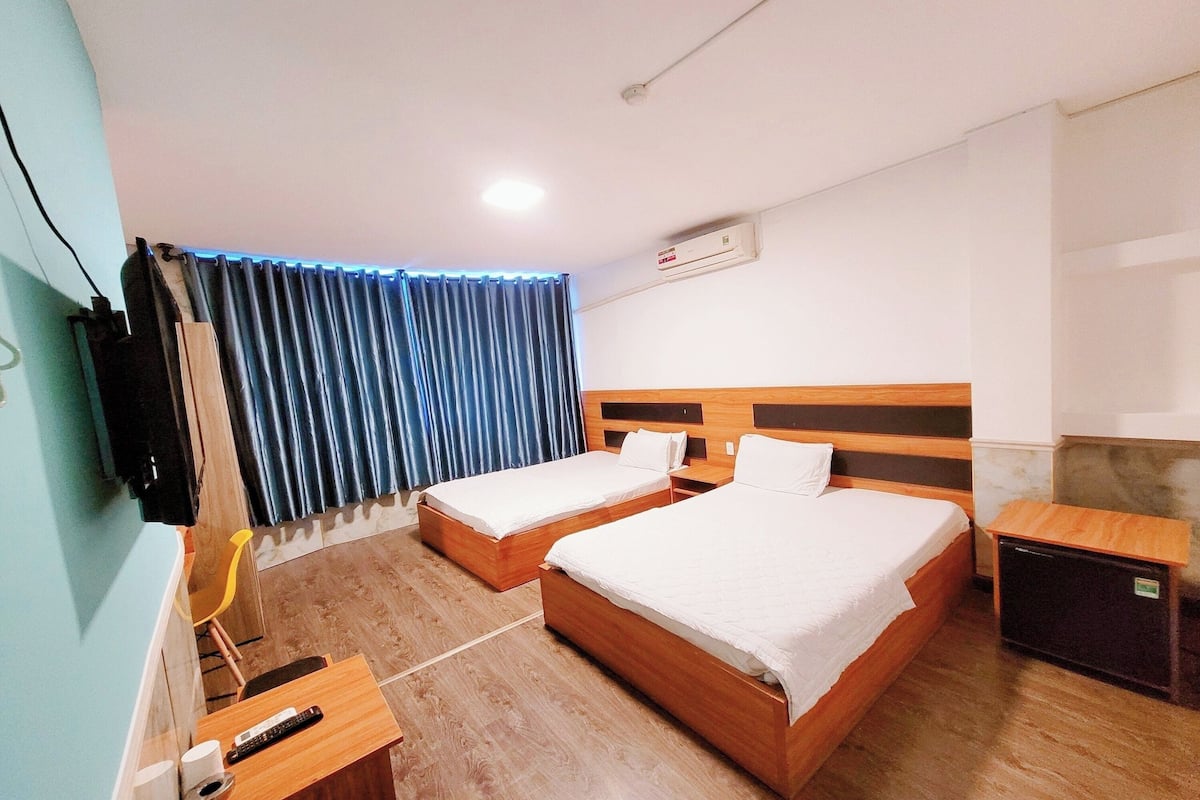 Une chambre d’hôtel avec deux lits, une tête de lit en bois, un petit bureau avec une chaise, une télévision et une fenêtre avec des rideaux bleus.