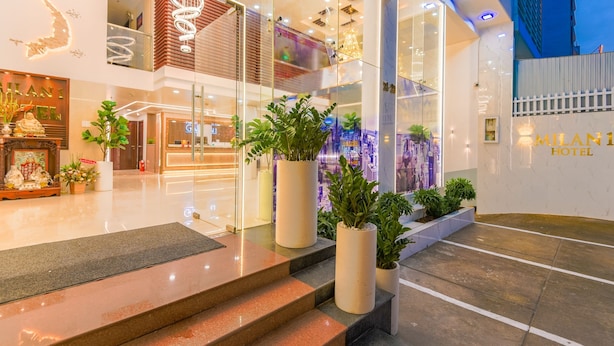 Un hall d’hôtel moderne avec une façade en verre, des plantes en pot et un escalier apparent menant à l’étage supérieur.