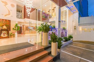 Um saguão de hotel moderno com fachada de vidro, plantas em vasos e uma escadaria visível que leva ao andar superior.