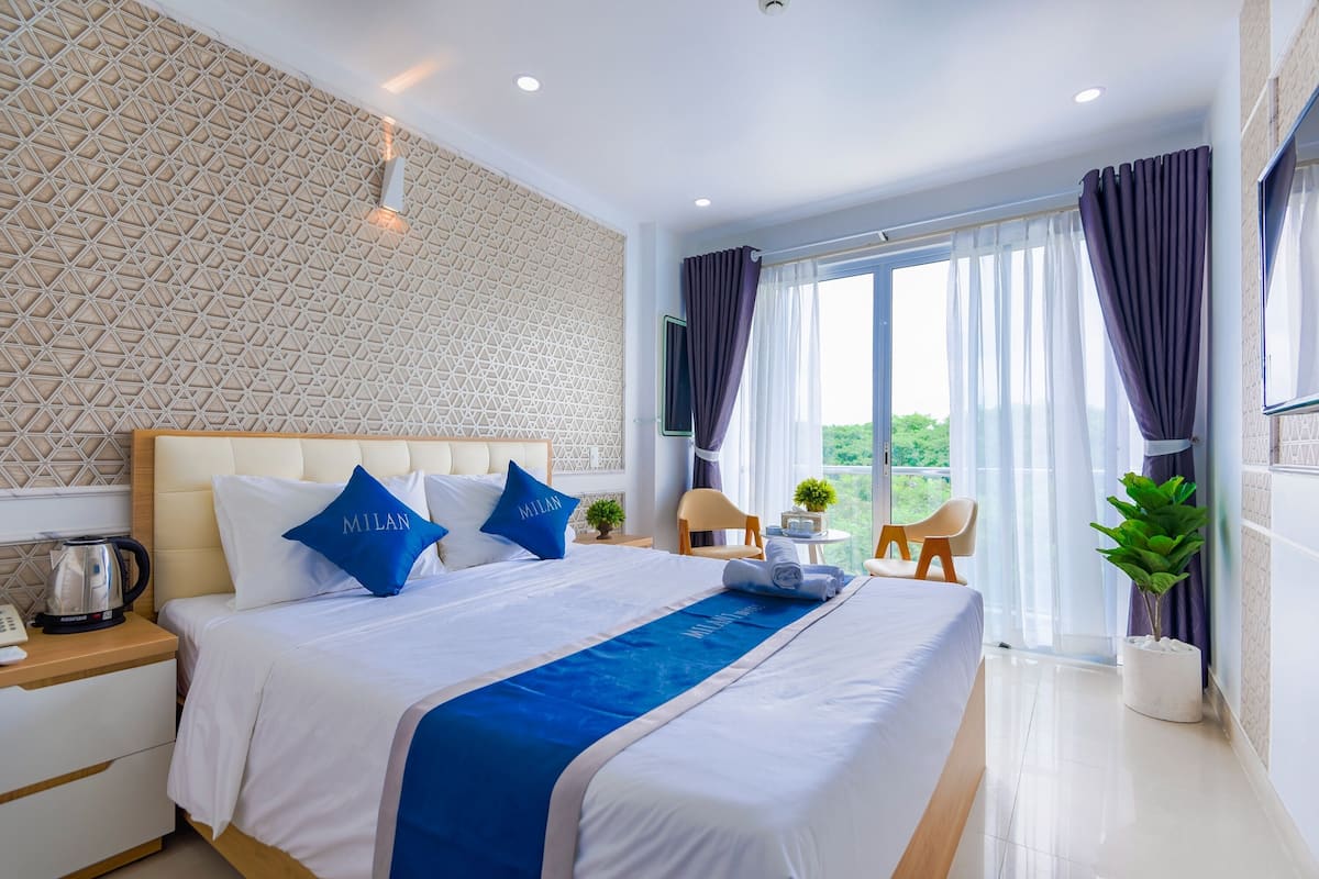 Une chambre d’hôtel avec un grand lit, une vue sur l’extérieur, une petite table avec une plante et une fenêtre avec des rideaux.
