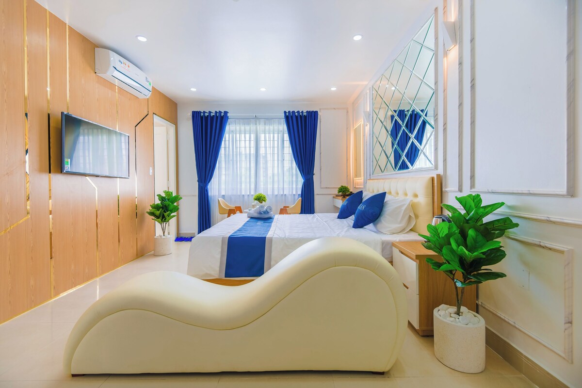 Une chambre d’hôtel moderne avec un grand lit, un téléviseur à écran plat, une plante en pot et une grande fenêtre ornée de rideaux bleus.