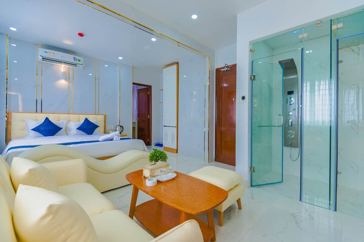 Une chambre d’hôtel moderne avec un lit, un canapé et une petite table. Elle comprend un espace douche fermé par des parois de verre et une porte donnant sur une autre pièce.