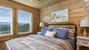 A bedroom with a wooden ceiling, a bed with a beige and white duvet, two bedside lamps, and a painting on the wall.