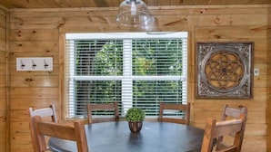 A wooden-paneled room with a round table, four chairs, a potted plant, a window with blinds, and a decorative metal piece on the wall.